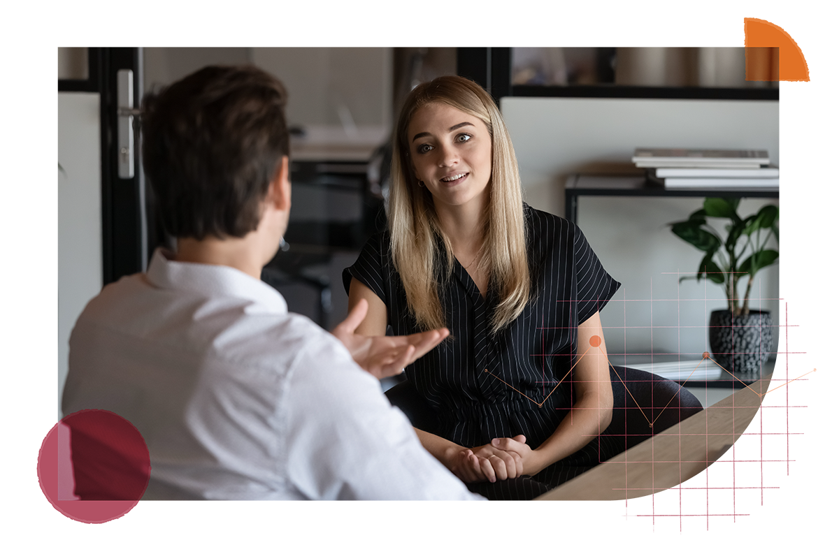 A gentleman interacting with a woman sitting at a desk. The gentleman is gesturing to the woman and is speaking with his back turned towards the camera. The woman has long blonde hair and is focused on what the gentleman is saying. She is wearing a black jumpsuit and they both appear to be in a open office area.